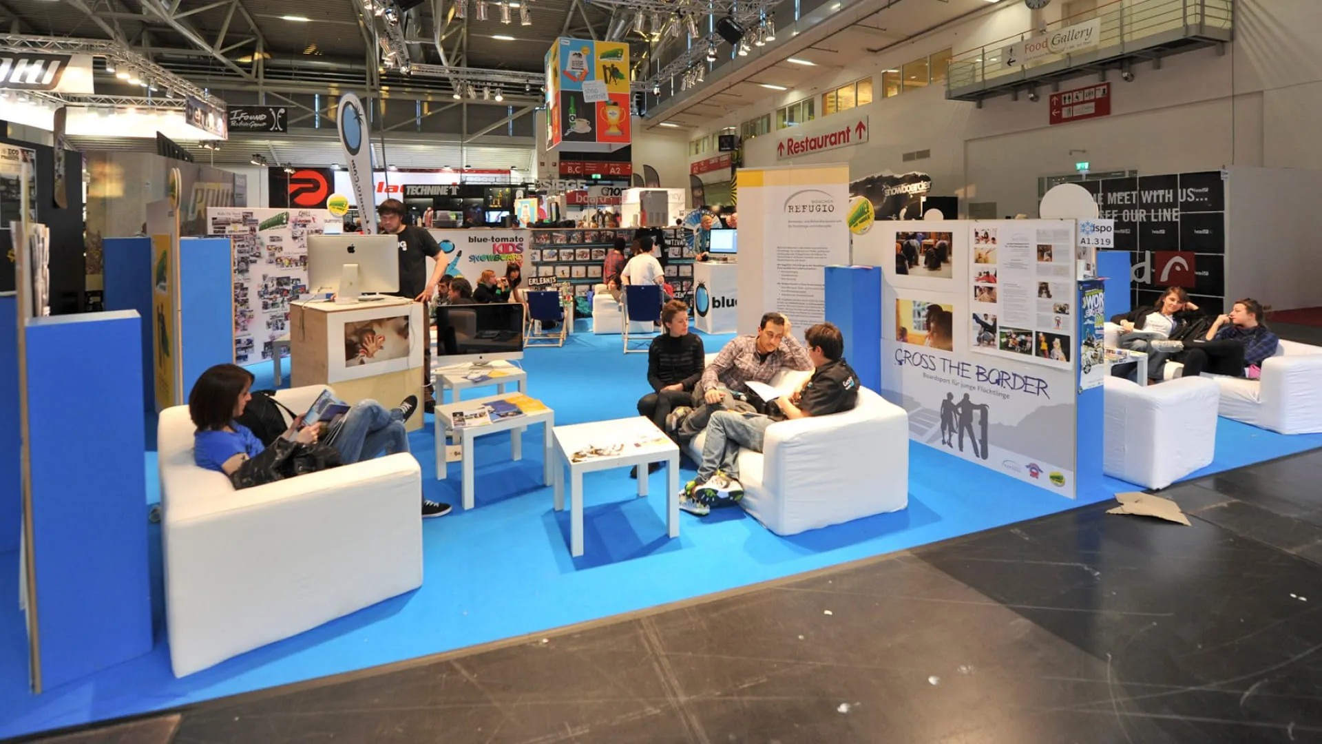 Messestand mit blauen Boden und weißen Sofas, Menschen sitzen